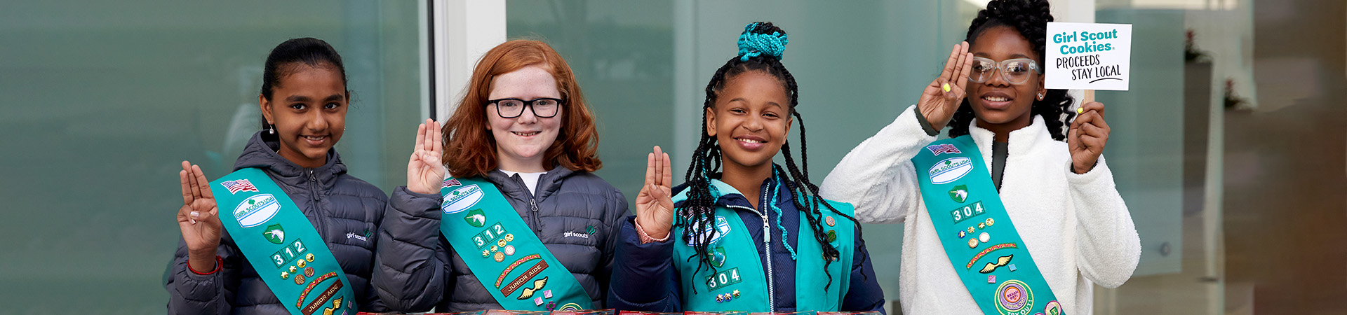  Four Girl Scout Juniors stand in front of a building holding a sign that says "Girl Scout Cookies Proceeds Stay Local" 