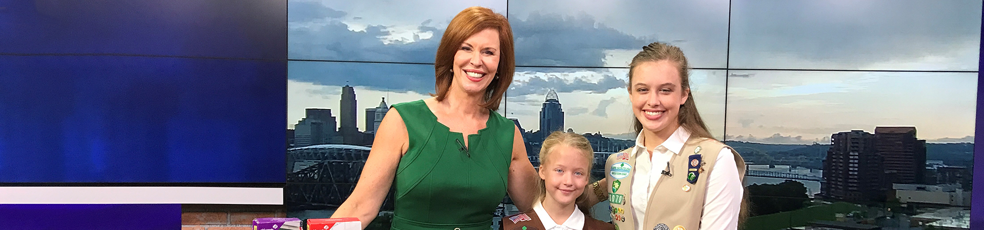 two girl scouts stand at a news desk with a newscaster 