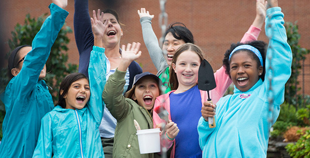 A group of girls and two volunteers holding gardening tools with their hands up cheering.